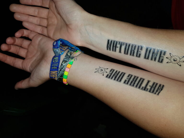 Nahaufnahme von Händen, die Tattoos mit der Aufschrift "Nature One" und bunte Festival-Armbänder tragen.