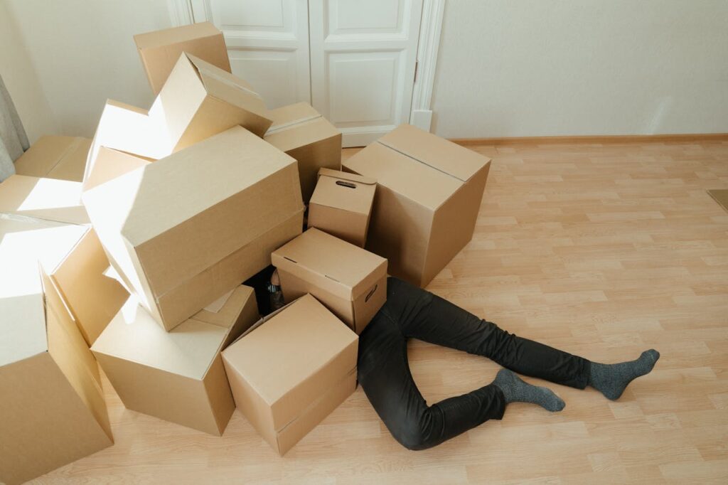 Eine Person liegt unter einer großen Menge brauner Umzugskartons, auf einem hellen Holzboden in einem Zimmer