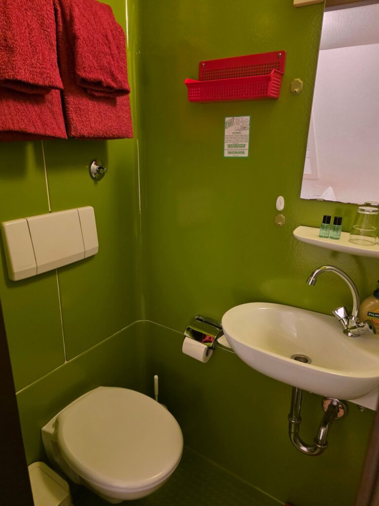 Modernes Badezimmer mit leuchtend grünen Fliesen, weißem Waschbecken, Toilette und roten Handtüchern. Kompakte Hotel-Badezimmer Einrichtung.
