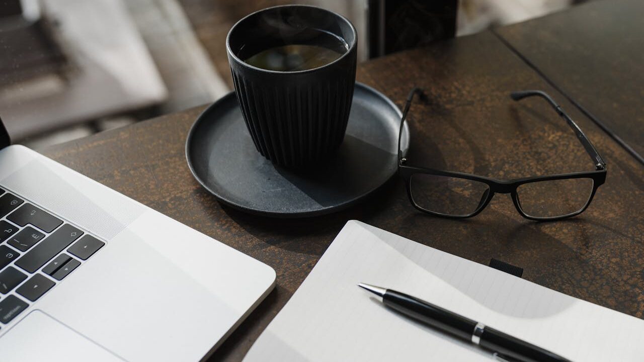 Blog erstellen als Anfänger (03.12.2025) Moderner Arbeitsplatz: Laptop, schwarze Kaffeetasse, einer Brille und Schreibmaterial auf dem Tisch