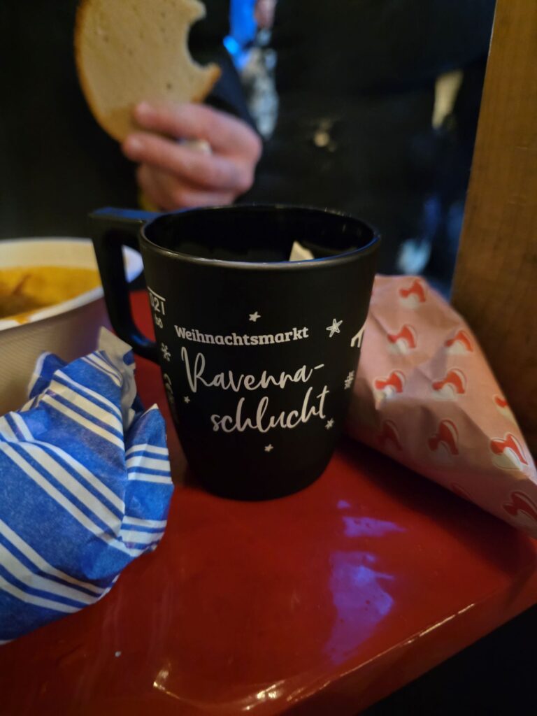 Schwarze Tasse mit weißem Schriftzug "Weihnachtsmarkt Ravenna Schlucht" auf einem roten Tablett. Daneben zwei Süßigkeitentüten