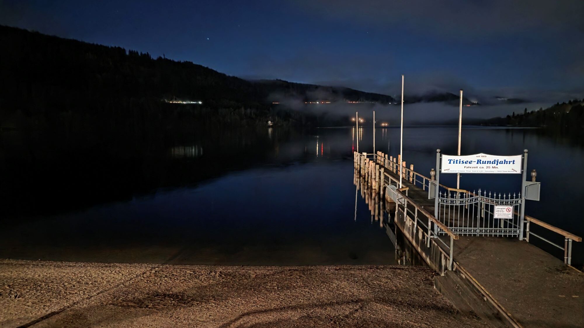 Beleuchteter Bootssteg am Titisee bei Nacht, mit ruhigem Wasser, bewaldeten Hügeln und einem Sandstrand im Vordergrund.