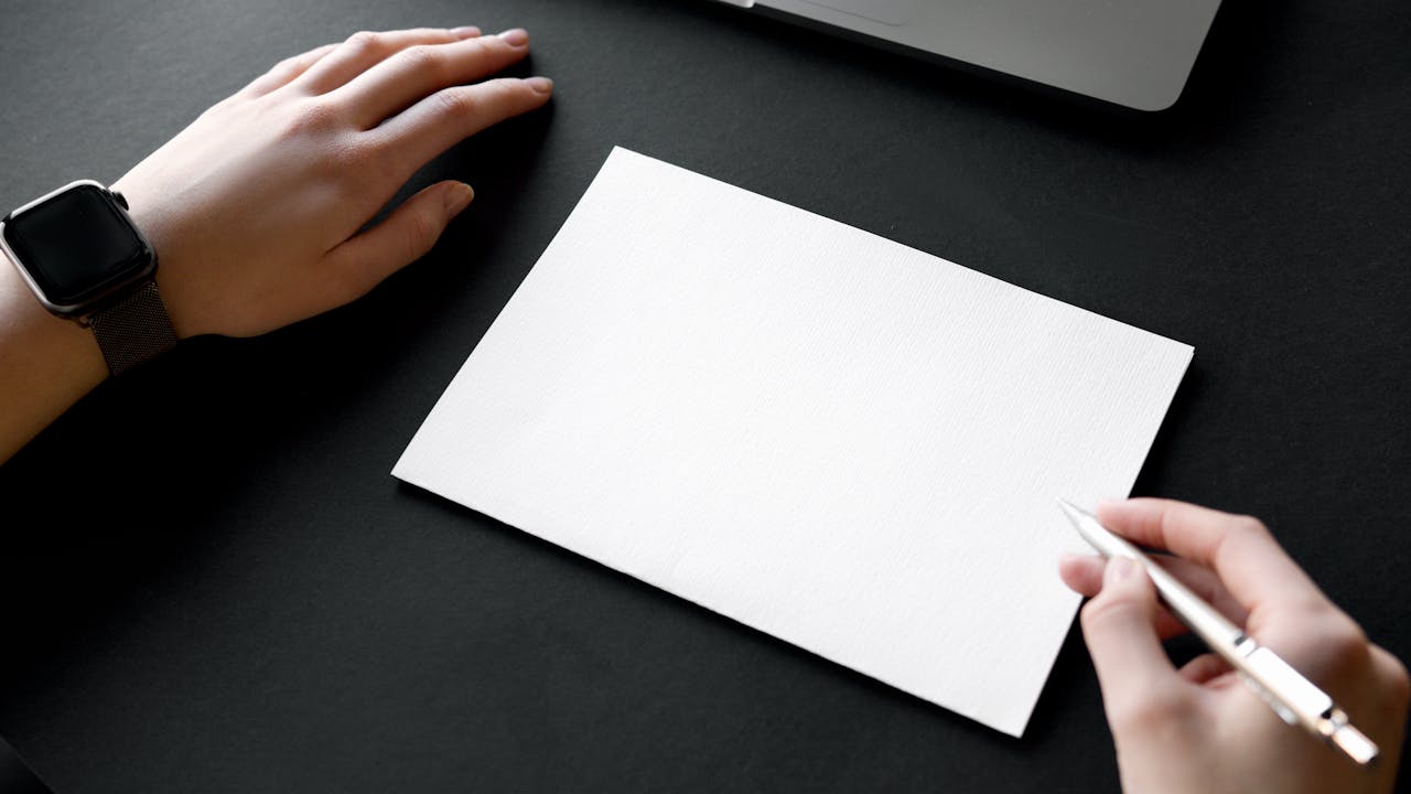 Blog erstellen als Anfänger (03.12.2025) Hände halten einen Stift über einem leeren Blatt Papier auf einem schwarzen Schreibtisch.