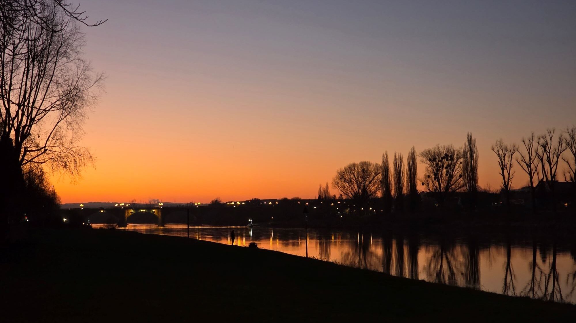 Silvester in Prag (04.01.2026) Flusslandschaft bei Sonnenuntergang mit Schattenrissen von Bäumen, einer Uferböschung und einer beleuchteten Brücke im Hintergrund