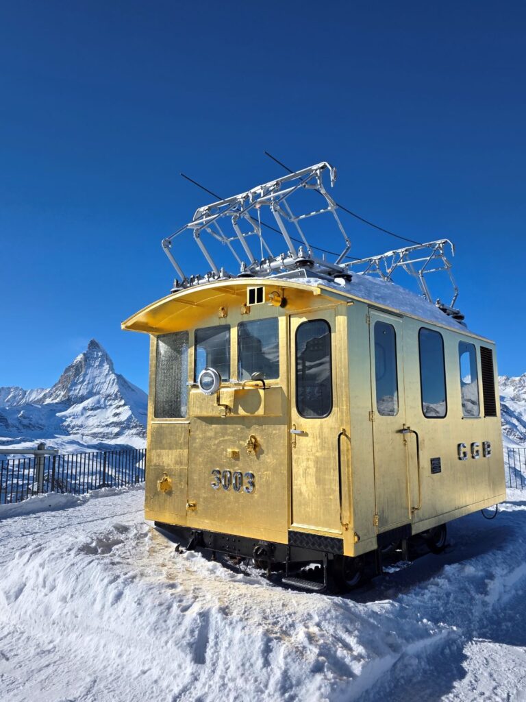 Glacier Express (01.02.2026) Eine ältere historische goldenen Lok auf einem verschneiten Plateau und im Hintergrund das Matterhorn
