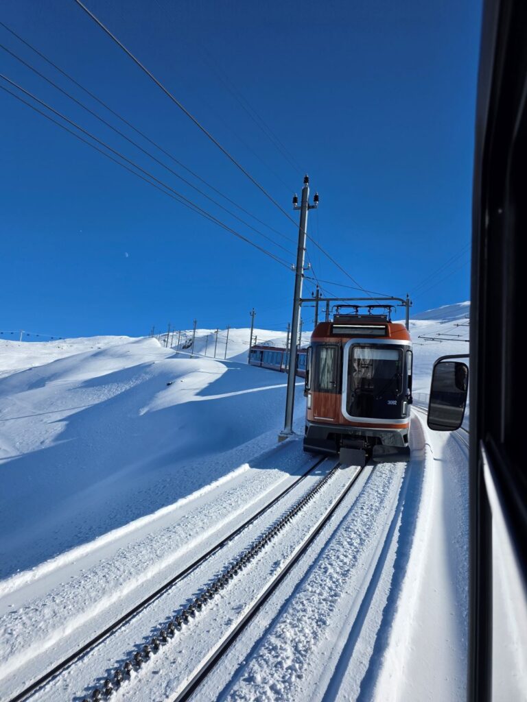 Glacier Express (01.02.2026) Blick aus einem Zugfenster auf eine entgegenkommende Gornergratbahn in verschneiter Winterlandschaft