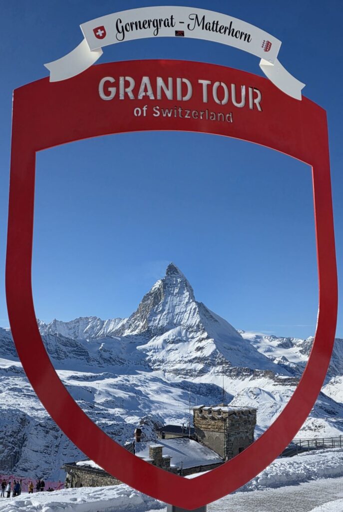 Glacier Express (01.02.2026) Blick durch ein rotes Schild mit der Überschrift "Grand Tour" auf das verschneite Matterhorn