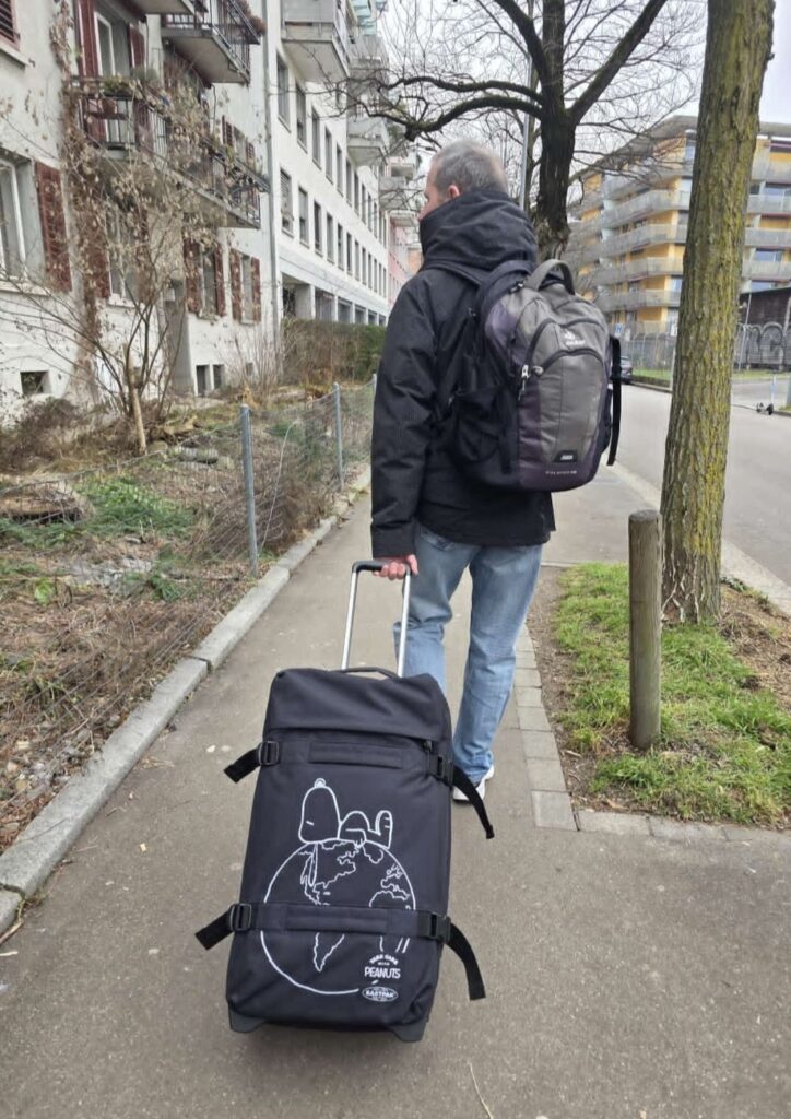 Glacier Express (01.02.2026) Mann in schwarzer Jacke von hinten, der einen Rucksack trägt und einen Koffer mit Snoopy Bild hinter sich herzieht