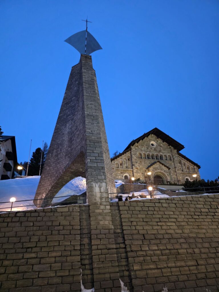 Glacier Express (01.02.2026) Eine Kirche im Hintergrund in der Dämmerung, davor ein hoher Turm mit einem Kreuz an dessen Spitze