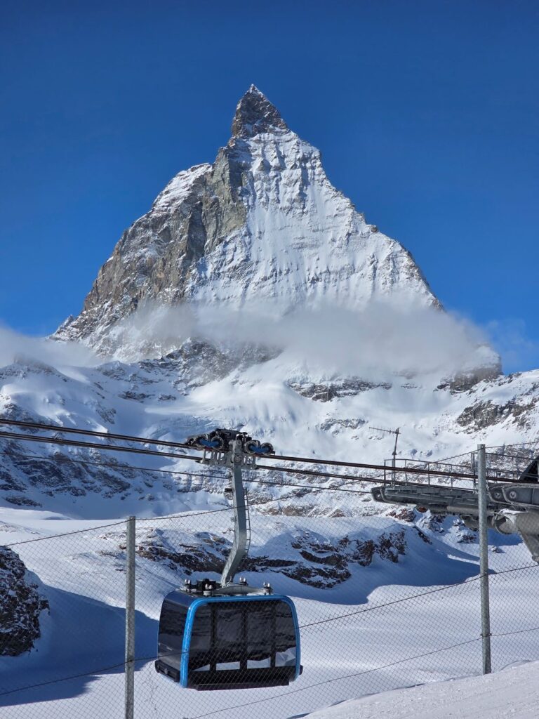 Glacier Express (01.02.2026) Verschneites Matterhorn im Hintergrund, im Vordergrund eine blaue Seilbahngondel während der Fahrt