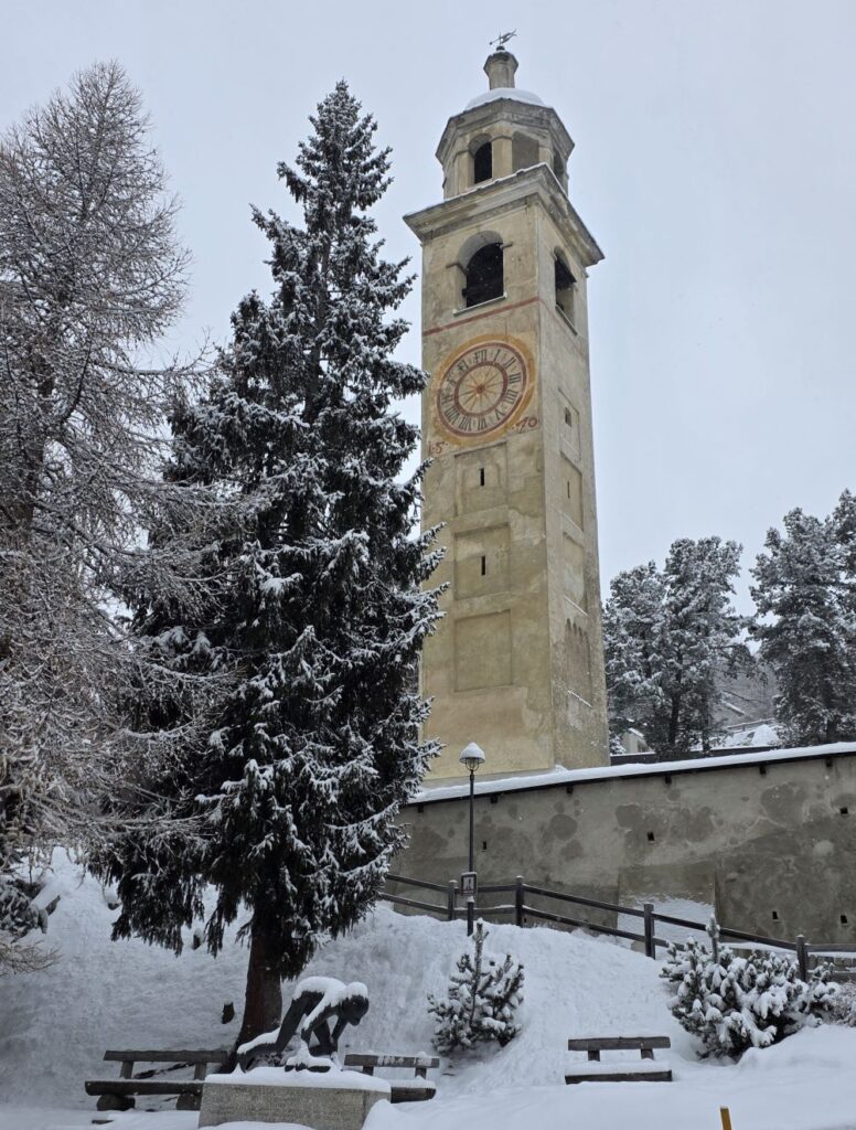 Glacier Express (01.02.2026) Schiefe Turm von St. Moritz einem alten Kirchturm in winterlicher Umgebung, davor verschneite Bäume und die Skulptur eines Bobfahrers