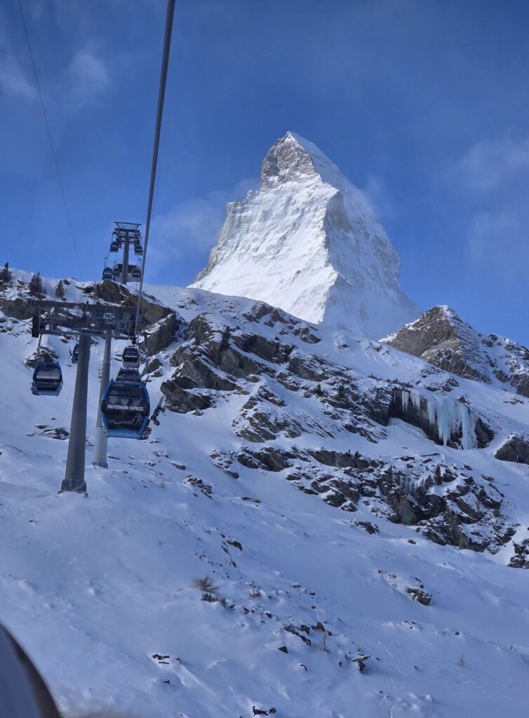 Glacier Express (01.02.2026) Blick aus einer Seilbahngondel auf davor fahrende blaue Gondeln bergauf und dem verschneiten Matterhorn im Hintergrund