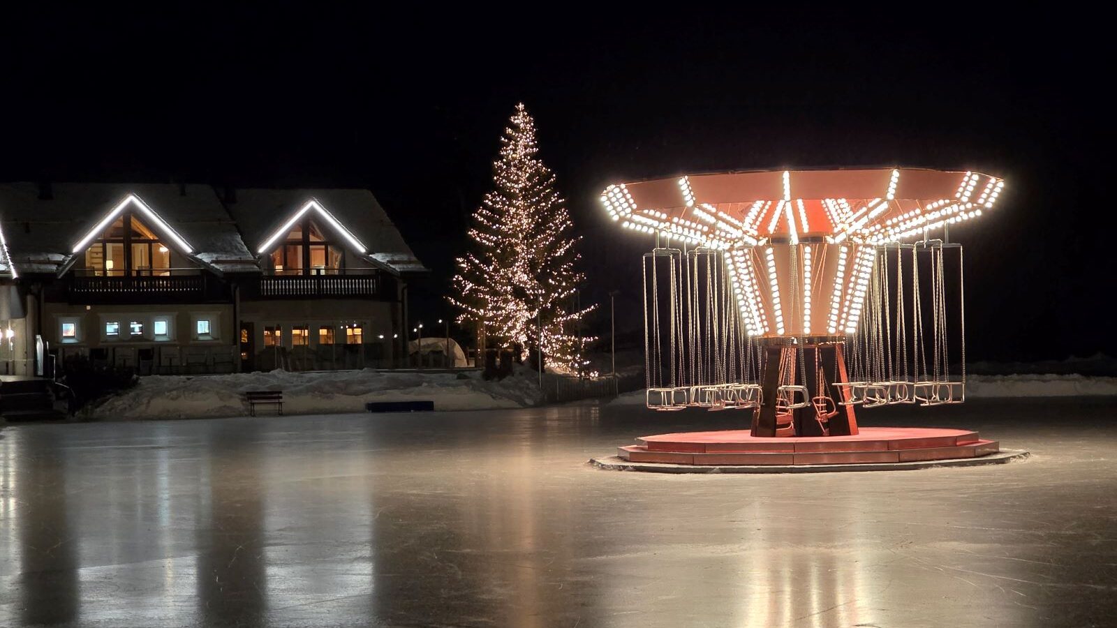 Glacier Express (01.02.2026) Eine Eislaufbahn in der Nacht mit einem hell erleuchteten Kettenkarrusell in der Mitte, einem beleuchteten Tannenbaum und beleuchteten Häusern im Hintergrund