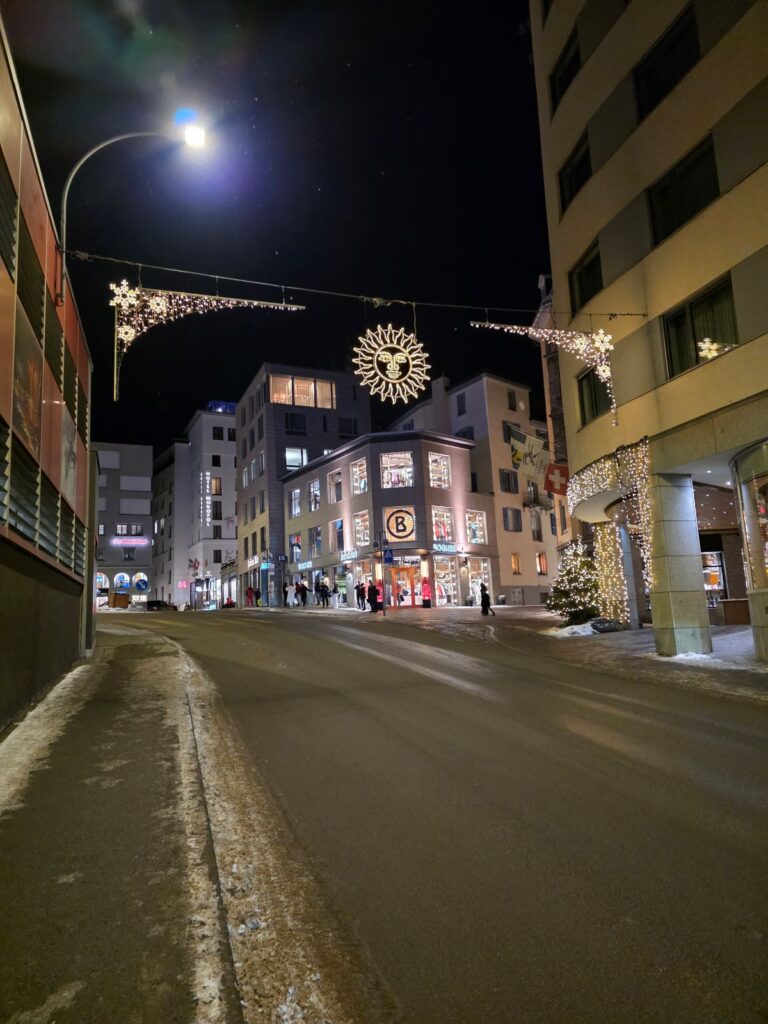 Glacier Express (01.02.2026) Straße in St. Moritz in der Nacht mit leuchtender Sonne über der Straße und beleuchteten Geschäften im Hintergrund