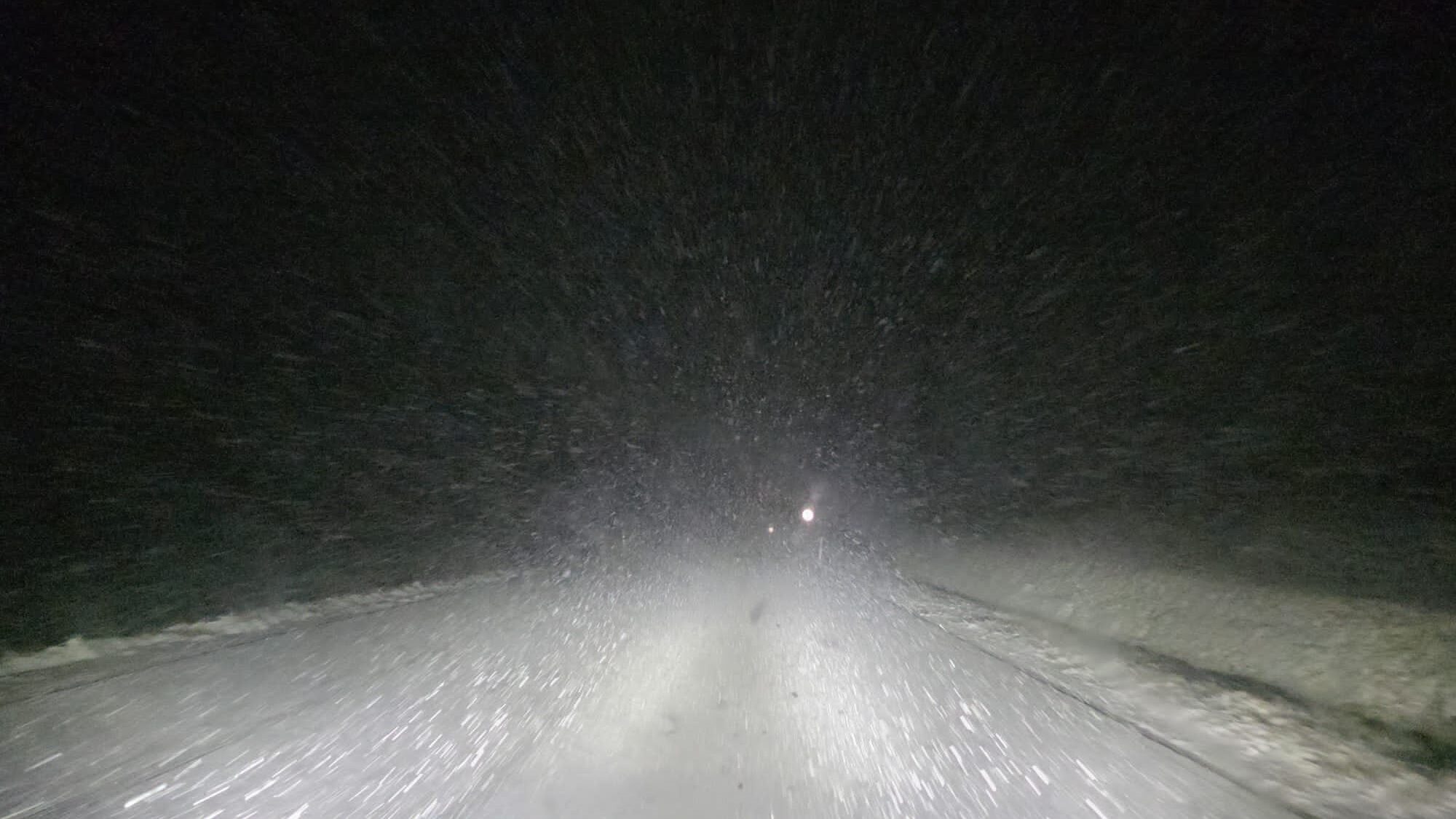 eine zugeschneite Strasse aus Sicht eines Autofahrers, es ist dunkel und die Schneeflocken erschweren die Sicht