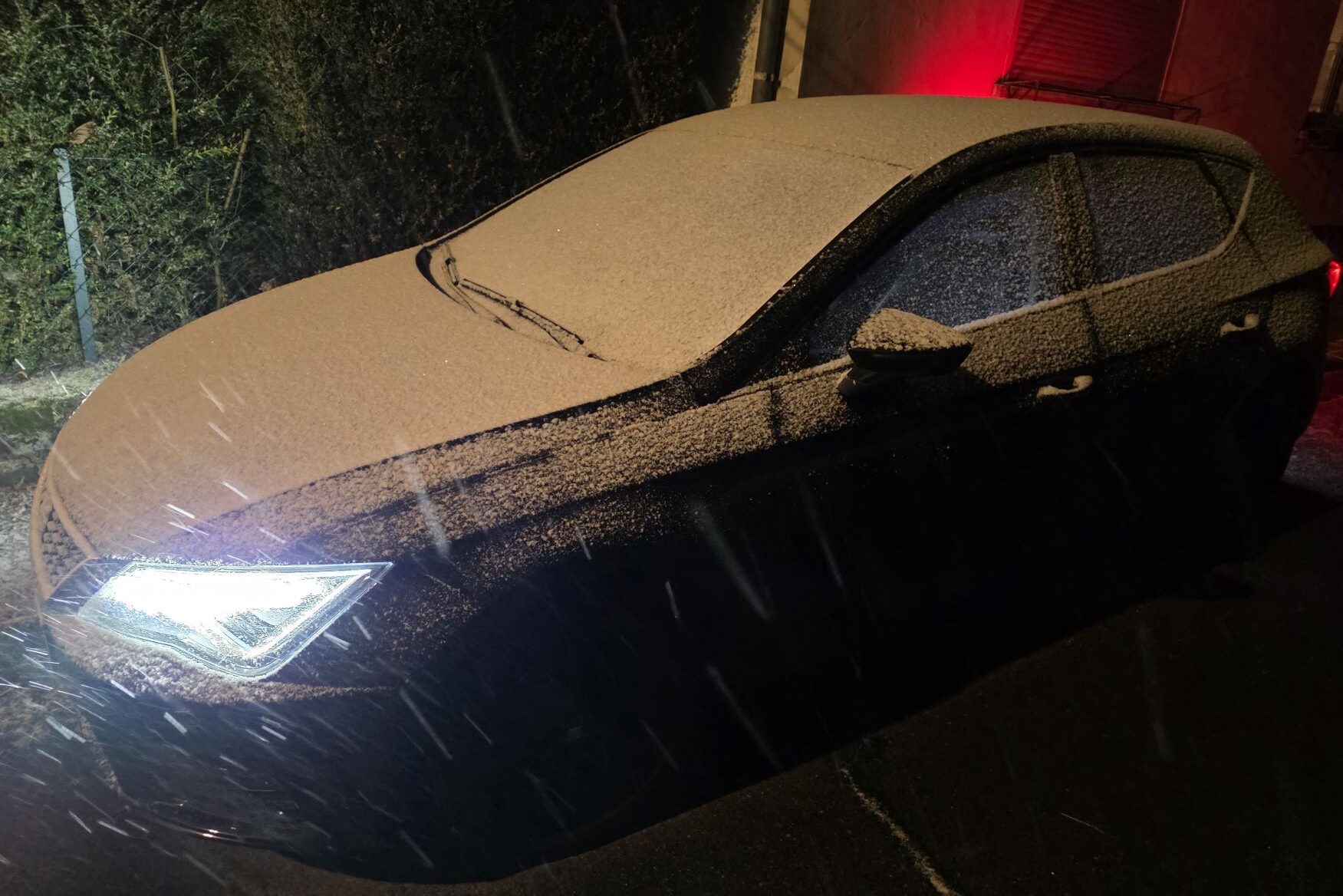Ein schwarzes Auto von der Seite steht vor einem Haus, auf dem eine dünne Schneeschicht liegt, es ist dunkel, es schneit und die Schweinwerfer sind an