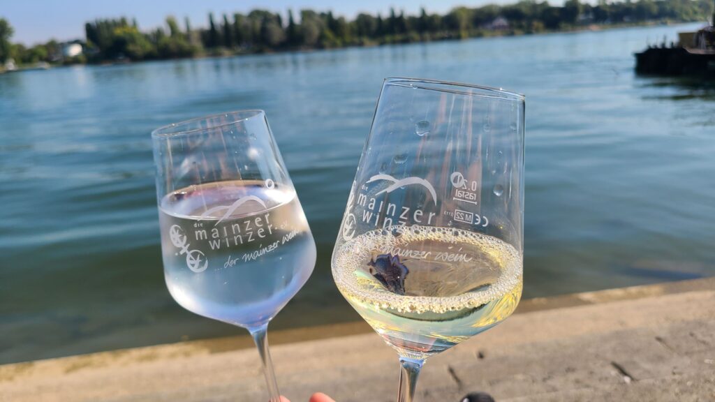 Stadtabenteuerspiel Mainz (13.02.2026) Zwei gefüllte Weingläser in die Kamera gehalten, dahinter der Rhein mit Baumreihen am anderen Ufer