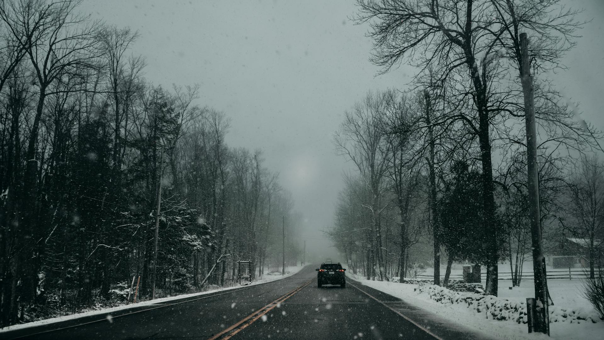 Home Straße bei plötzlichem Wintereinbruch auf der ein Auto fährt, es schneit und am Straßenrand stehen verschneite Bäume