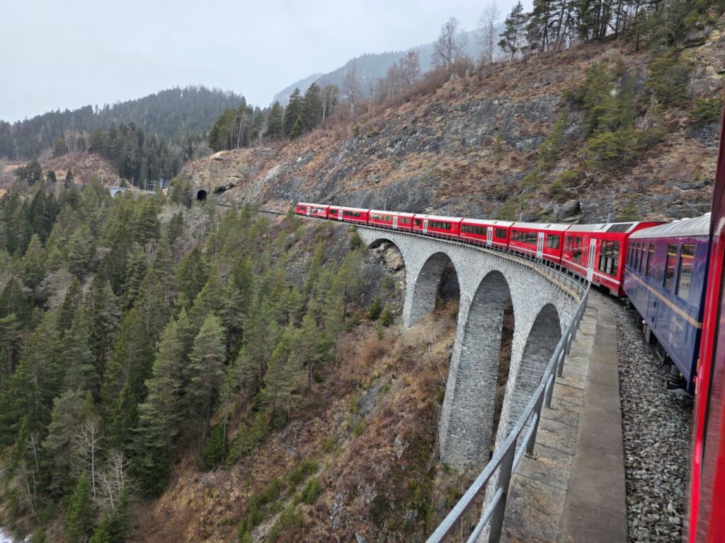 Glacier Express (01.02.2026) Blick aus einem Zugfenster auf den gesamten Zug von der Seite wie dieser über eine hohe Brücke fährt