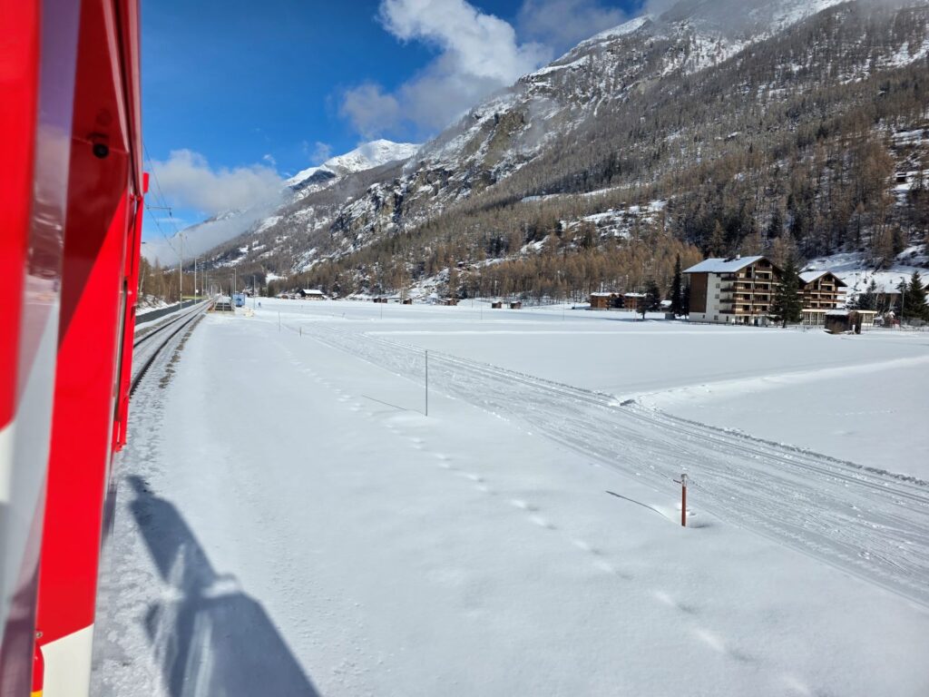 Glacier Express (01.02.2026) Blick aus dem Zug auf eine verschneite Winterlandschaft mit Wolken und hohen Bergen im Hintergrund