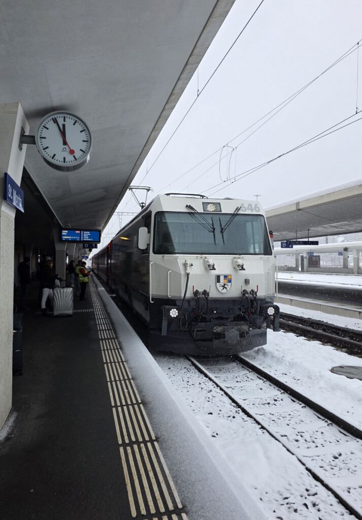 Glacier Express (01.02.2026) Bahnsteig mit einer Uhr an der Decke, sowie einer weißen Lok von vorne und den Wagons dahinter