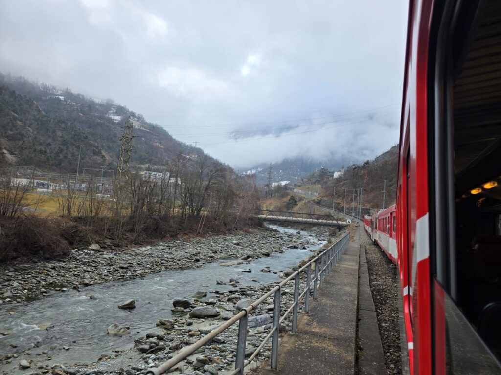 Glacier Express (01.02.2026) Blick aus einem Zugfenster auf einen fahrenden Zug an einem kargen Fluß vorbei. Im Hintergrund sind einzelne Häuser durch Bäume zu erkennen